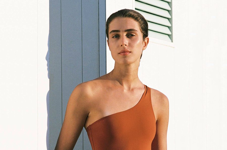 A campaign portrait: a model in a terracotta one-shoulder swimsuit against a pale shuttered wall in hard sunlight.